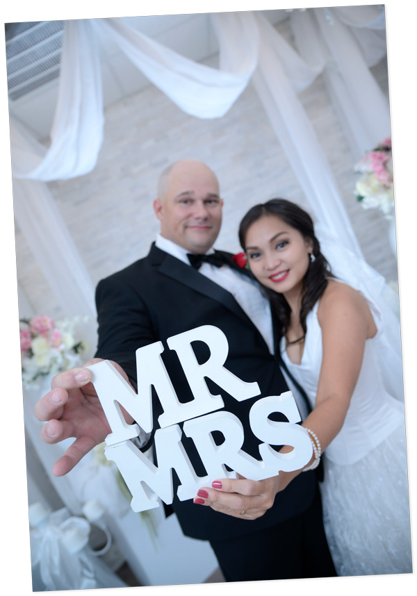 In a joyous moment at Lucky Little Chapel, this image features a newly married couple standing together, both dressed in formal attire. The man holds a large white sign reading 'MR & MRS,' while the woman embraces him. Surrounded by potted plants and a vase, the couple's happiness shines through as they commemorate their special day.